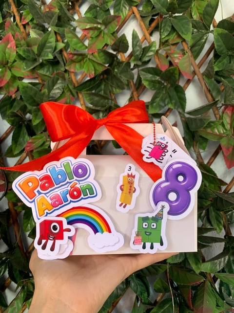 Hand holding a white gift box decorated with colorful Papilaon stickers and a red ribbon bow, with green ivy vines in the background