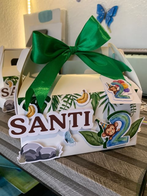 White decorative gift box with tropical fruit and monkey designs, green ribbon bow, and SANTI text label on wooden surface
