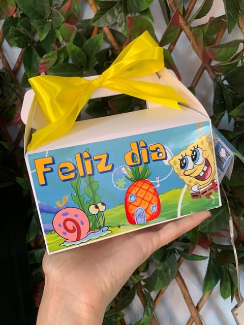 Hand holding a white gift box with Feliz dia message and cartoon characters, topped with a yellow bow against greenery