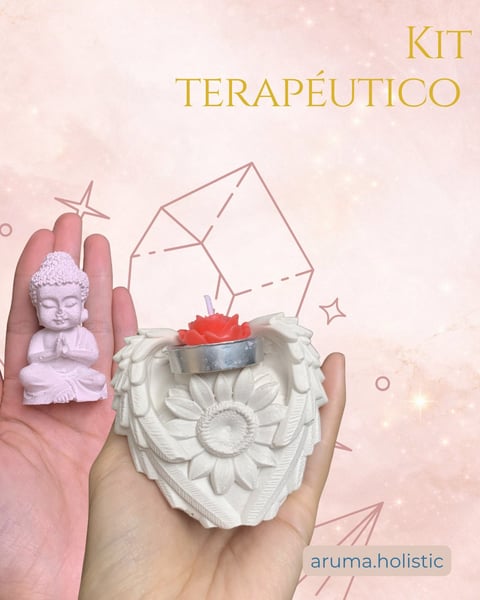 Hands holding decorative items: a small white Buddha figurine and a white flower-shaped holder with a red candle. Text reads KIT TERAPÉUTICO with aruma.holistic logo on soft pink background